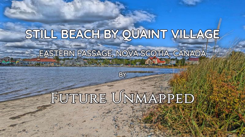 Sandy beach curves towards idyllic, tranquil fishing village - with red-roofed church, parked vehicles, and tourist area - running alongside large-stoned shoreline.